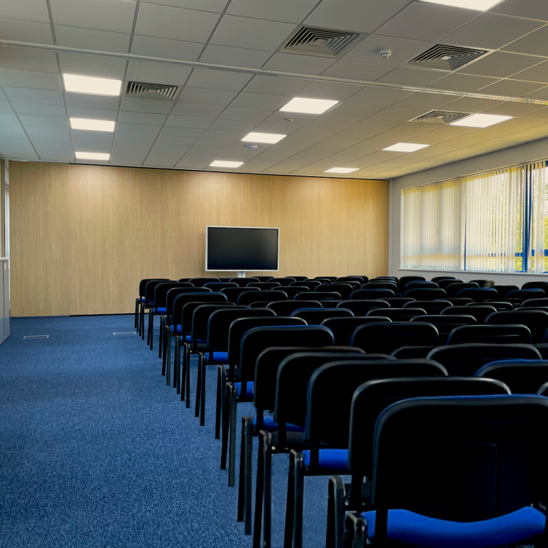 A photo of a large theatre layout meeting room and large screen at the front against the pine effect wall.