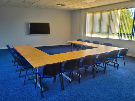 An empty training room with a u-shape layout of tables with 14 chairs around them, with a 65" smart screen on the wall