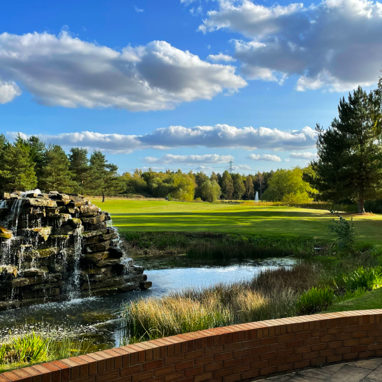 A photo of golf course on a sunny day with a water fountain, trees, and blue skies.