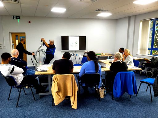 A photo of a training room in use. Students are sitting watching a demonstration of a hoist being used.