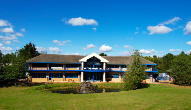 A photo of a building with grass and a water fountain in front of it, to the sides are nice green trees and there is bright blue sky.