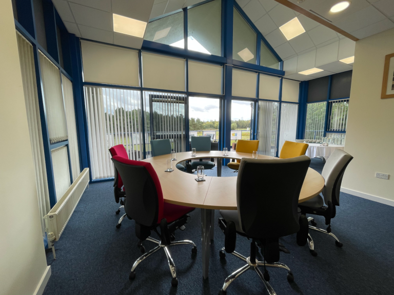 A photo of a boardroom with a circular table and eight colourful chairs around it.