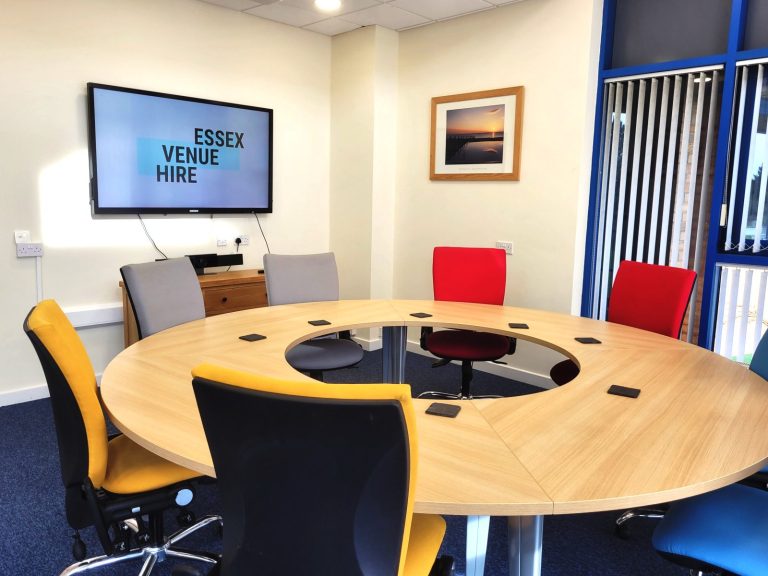 A photo of the round table in the boardroom with colourful, cushioned chairs around it, and the Essex Venue Hire logo on the 64" smart TV