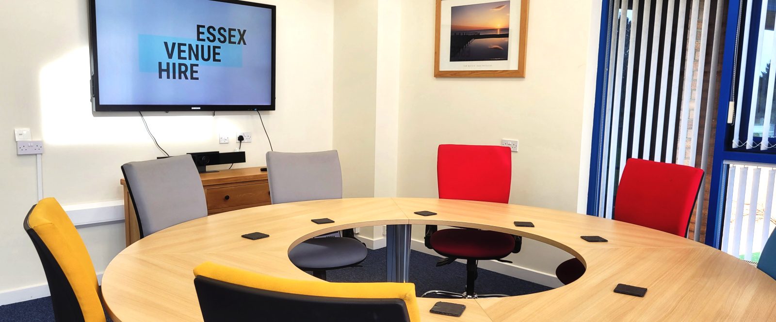 A photo of the round table in the boardroom with colourful, cushioned chairs around it, and the Essex Venue Hire logo on the 64" smart TV