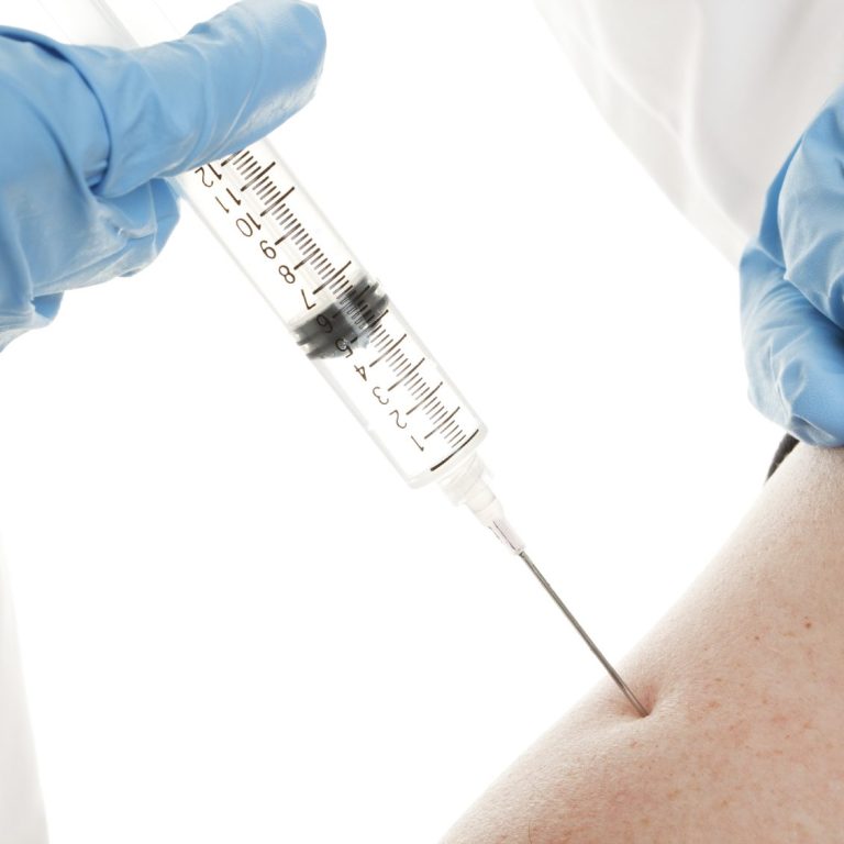 A photo of a syringe in an arm with a blue gloved nurses hand on the plunger