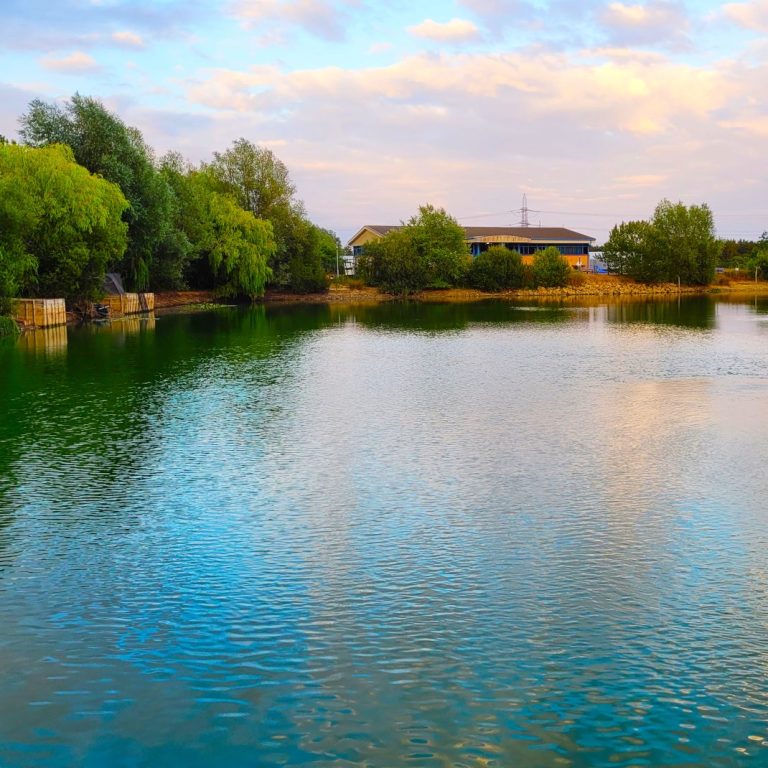 A photo of Earls Colne Fishing lake with tranquil water, surrounded by trees and an office building in the background with blue sky and clouds