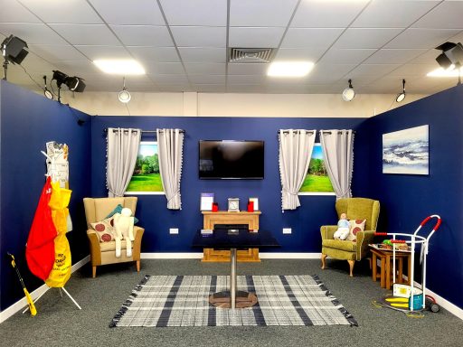 A photo of a studio set of a living room with arm chairs, a table, a TV on the wall, and a rug on the floor.