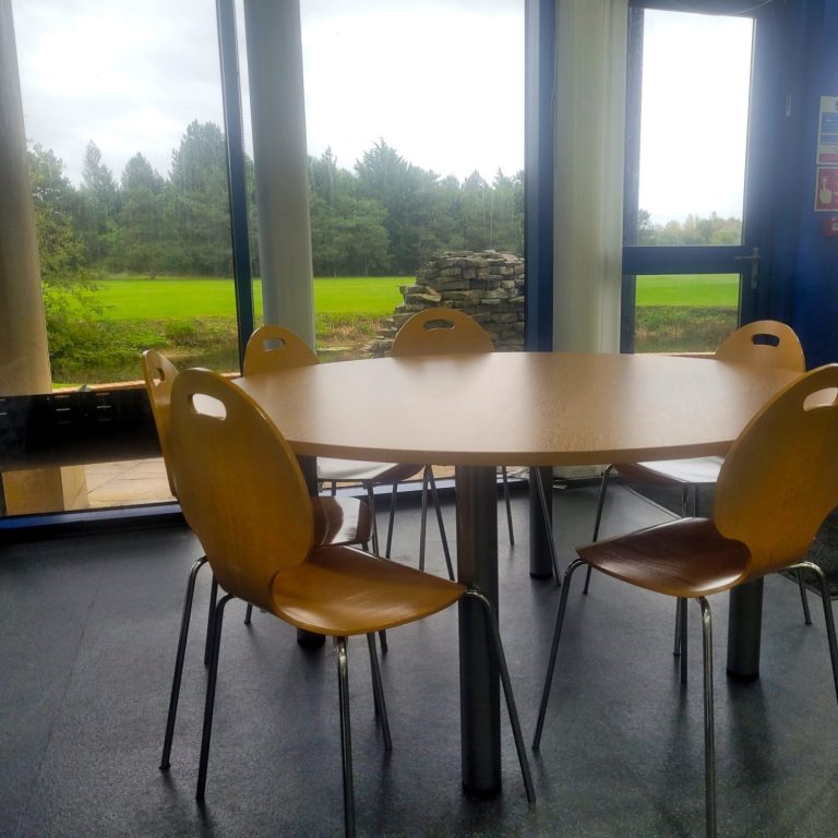 A photo of a pine circular table and matching chairs in a staffroom, with views of a golf course through the windows