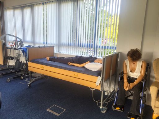 A photo of training equipment, a profile bed with mannequin laying on it and a wheelchair and mannequin sitting in it.