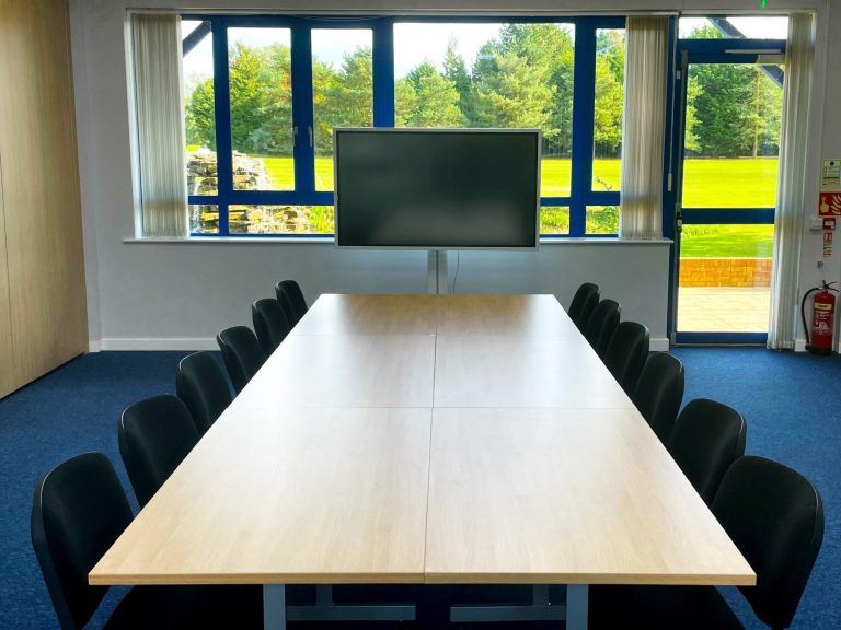 A photo of a meeting room with a boardroom layout and 64" smart TV at the head of the table and the views out the window behind it