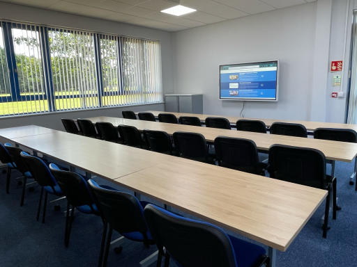 A meeting room with classroom layout, 3 rows of tables and chairs, and large interactive smart screen on the wall with website displayed.